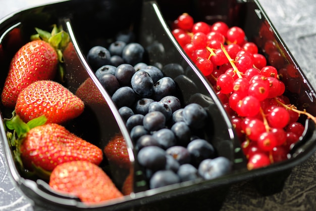 Recipiente de plástico con bayas, grosellas rojas, moras y fresas en el fondo gris