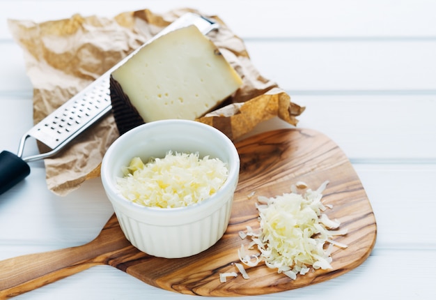 Recipiente con queso rallado sobre una tabla de cortar. Pedazo de queso y rallador. Copie el espacio.