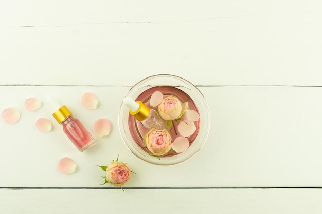 Recipiente de vidrio con agua de rosas y pétalos de rosa, dos botellas de aceite de rosa cosmético sobre un fondo de madera blanca. vista superior.