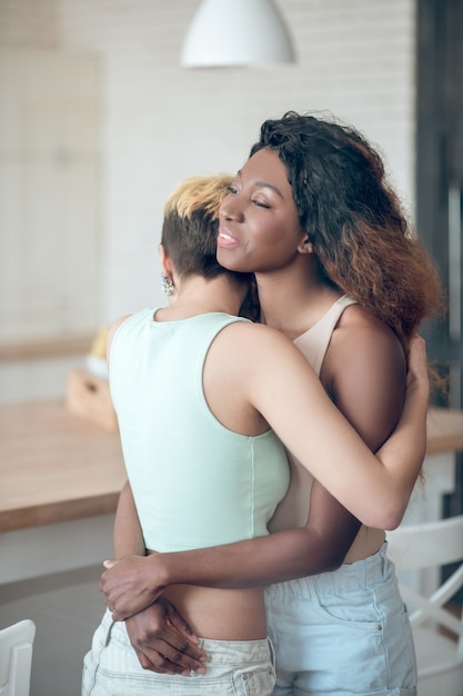 Reciprocidad. Dos amigas delgadas adultas jóvenes en ropa casual de pie cerca uno del otro abrazándose en casa