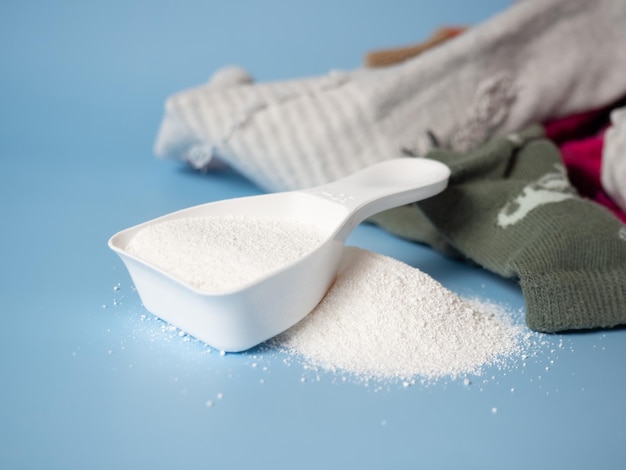 Recogedor blanco con detergente en polvo para bebés en el fondo de la ropa Detergente para lavar la ropa de los niños Limpieza en seco Lavado regular Espacio en blanco para el texto sobre un fondo azul pastel