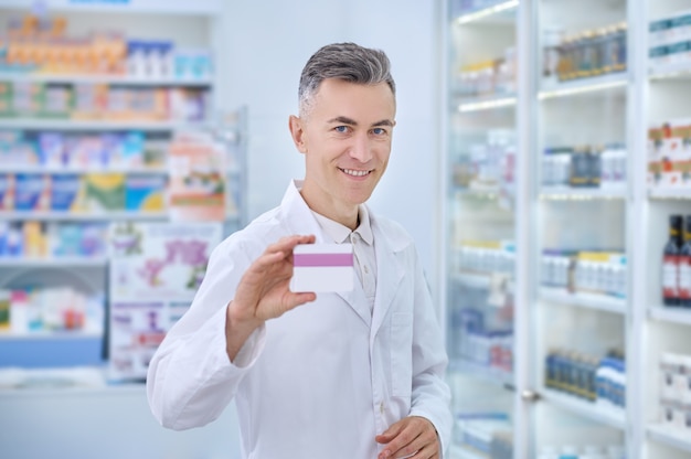 Recomendação. homem adulto de jaleco branco sorrindo com otimismo e mostrando medicamento em pé perto de prateleiras com remédios na farmácia