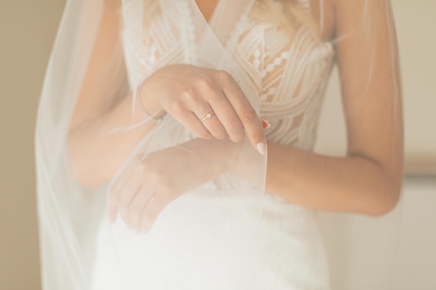 Recorta la foto de las manos irreconocibles de la novia con manicura ligera, anillo y pulsera, usando un vestido de novia blanco y un velo de novia mientras se prepara para el día de su boda. reunión de la mañana de la novia