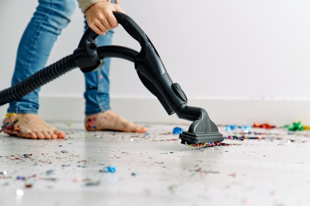 Recorta a un niño anónimo descalzo con jeans azules usando una aspiradora para recolectar confeti colorido del piso después de la fiesta