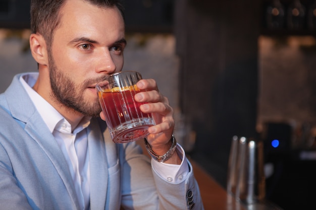 Recortada cerca de un apuesto hombre elegante con barba bebiendo cóctel de whisky en el bar