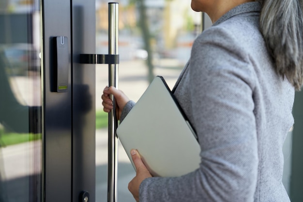 Recortada por debajo de una mujer de negocios sosteniendo una computadora portátil y abriendo la puerta principal de la oficina