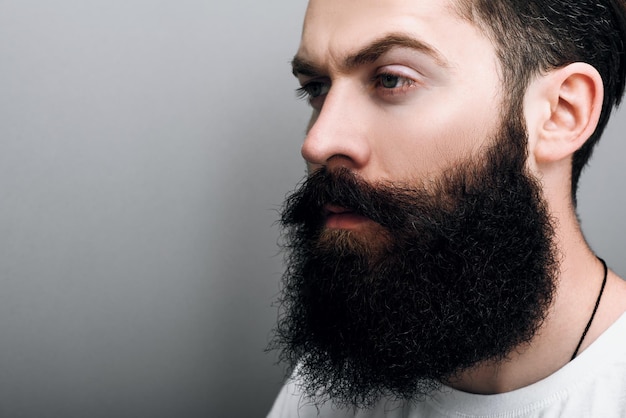 Recortado close-up retrato de homem caucasiano bonito brutal com barba na moda e bigode posando no fundo do estúdio cinza Barbeiro barbudo modelo de homem europeu com expressão confiante no rosto