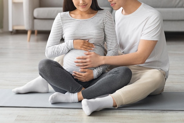 Recortado de jovem esperando família curtindo aula de fitness juntos, marido e mulher grávida irreconhecível sentado no tapete de ioga, abraçando a barriga grande da mulher, espaço de cópia, interior de casa