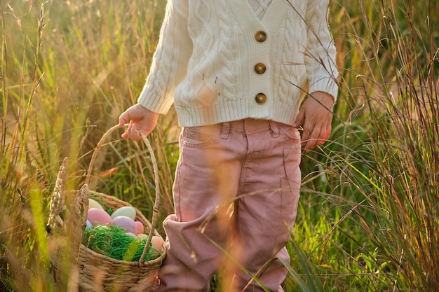 Recortar a una chica irreconocible con suéter blanco que lleva coloridos huevos de Pascua en una canasta de mimbre mientras está de pie en el césped en un campo soleado