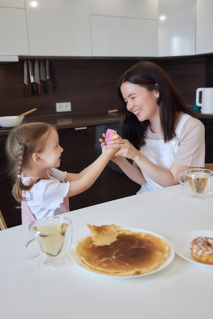 Recortar foto de madre en rojo y su hija con donuts coloridos sentados en la cocina Concepto de dieta y comida chatarra