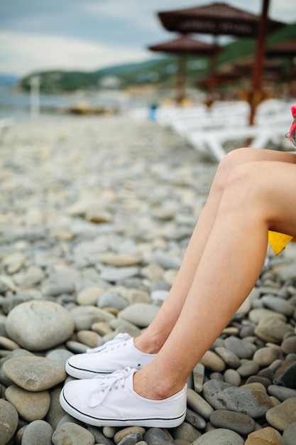 Recortar foto de mujer con las piernas desnudas usando zapatillas blancas y posando en la playa de guijarros