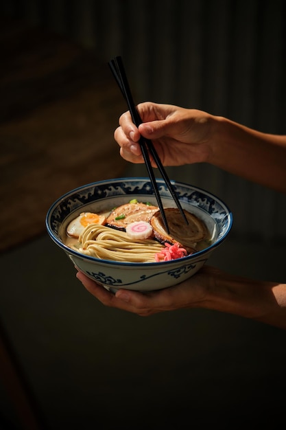 Recortar mujer anónima con palillos comiendo deliciosa sopa de ramen con fideos y carne en un tazón sobre fondo oscuro