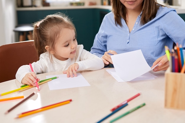 Recorte a mãe amorosa e a adorável filha sentadas à mesa com lápis coloridos e folhas de papel enquanto desenham juntas durante o fim de semana em casa