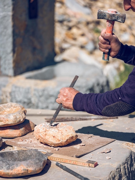 Recorte a un artesano anónimo cortando roca usando cincel y martillo mientras crea artesanías de piedra en un banco de trabajo en un día soleado