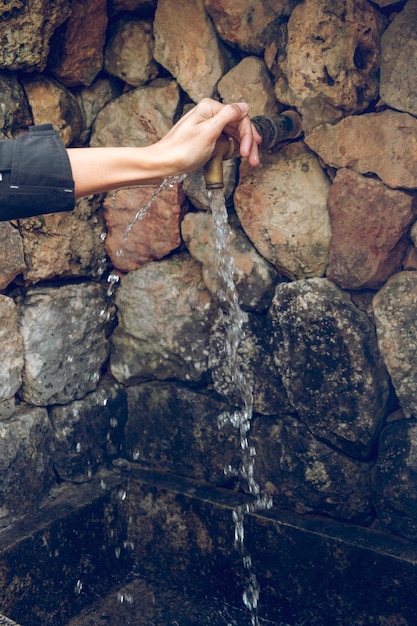 Recorte a una mujer sin rostro tirando de un grifo de cobre en una antigua pared de piedra mientras abre agua mineral pura durante el día