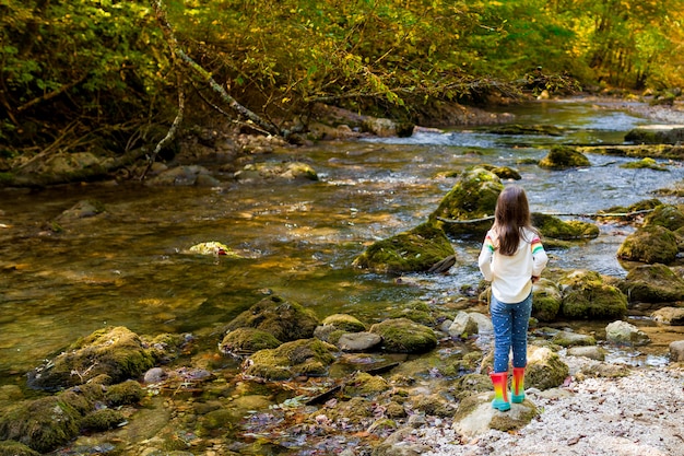 Recreação ao ar livre e incríveis aventuras com crianças. uma menina criança está andando ao longo de um rio verde na floresta em botas de borracha em um dia quente de outono