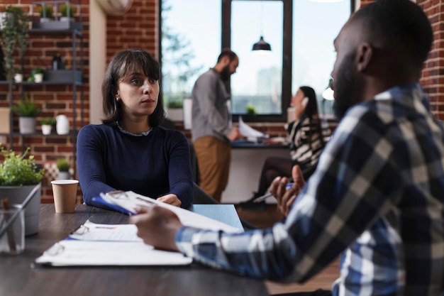 Recrutador de empresa de negócios e candidato sentado na mesa no escritório enquanto revisa o currículo do candidato a emprego. Pessoas multiétnicas na agência de startups falando sobre oferta salarial e regulamentos internos.