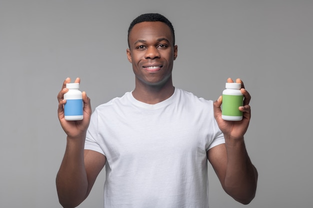 Recuperação. Homem alegre e atraente de pele escura segurando, mostrando pacotes de vitaminas nas mãos, em pé no estúdio