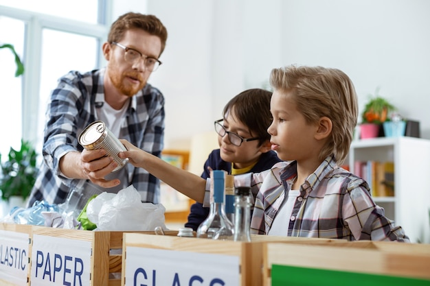 Recycling-System. Neugieriger blonder Junge, der Müll in die richtigen Kisten legt, während er eine pädagogische Übungsstunde hat