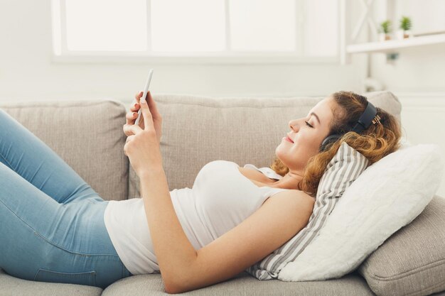 Redes sociais. Mulher jovem ruiva enviando mensagens no smartphone em casa, deitada confortavelmente no sofá bege, copie o espaço