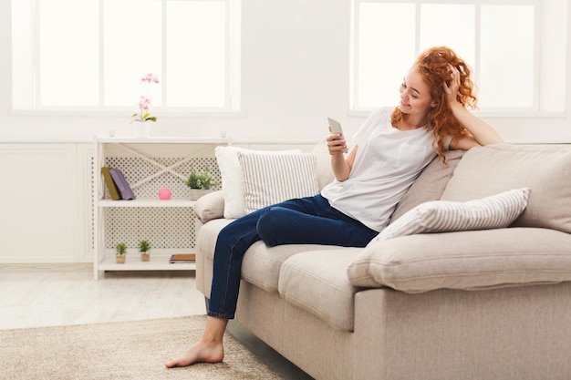 Redes sociales. Mujer joven pelirroja enviando mensajes en el teléfono inteligente en casa, sentada cómodamente en el sofá beige, espacio de copia