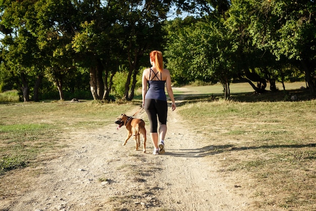 Redhair jouful joven acariciando a su perro usando ropa deportiva disfrutando de su tiempo y vacaciones en el parque soleado