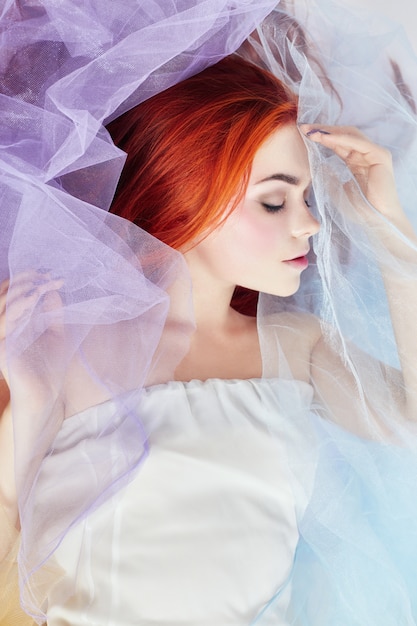 Redhead Girl dreams, maquillaje brillante, piel limpia, cuidado facial. Chica pelirroja en un vestido de color claro se encuentra en el suelo, un retrato de primer plano. Romántica mujer con cabello largo y vestido de nube