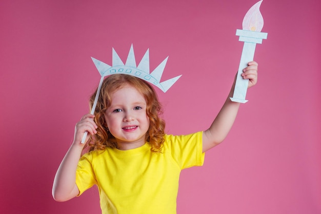 Redhead Ingwer charmante weibliche Kinderfeier Unabhängigkeit hält eine Papierfackel und DIY-Krone auf einem rosa Hintergrund im StudioEnglisches Sprachlernkonzept und Freiheit