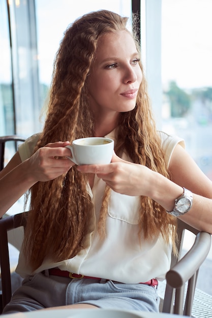 Redhead lockiges weißes dünnes Mädchen im weißen Hemd, das Kaffee im Café trinkt und Fenster betrachtet