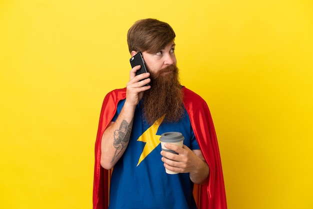 Redhead Super Hero Mann isoliert auf gelbem Hintergrund mit Kaffee zum Mitnehmen und einem Handy