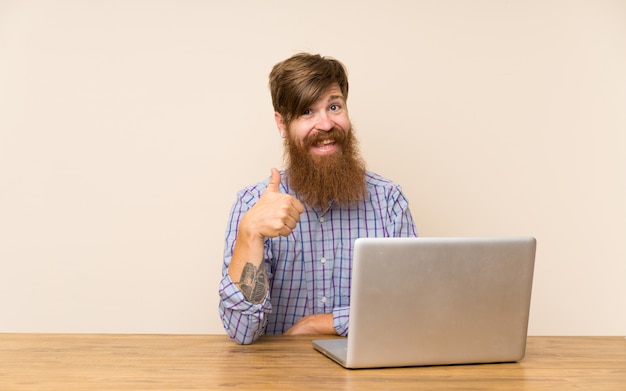 Redheadmann mit langem Bart in einer Tabelle mit einem Laptop, der Daumen gibt, up Geste