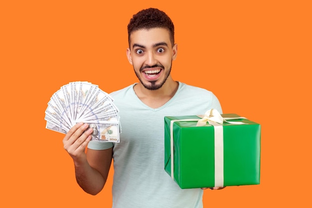 Reembolso de compras. Retrato de un hombre extremadamente feliz con barba en pantalones blanco sosteniendo una caja de regalo y mucho dinero, mirando con asombro, el cliente gratamente sorprendido con las bonificaciones. tiro de estudio interior