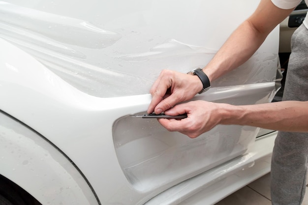 Reequipando el coche con una película protectora transparente sólida, el maestro hace una incisión con un cuchillo en el revestimiento que protege los vehículos de arañazos y astillas en el taller