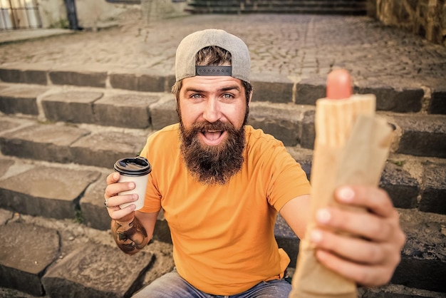 Refeição com gosto certo Homem barbudo comendo alimentos pouco saudáveis durante o descanso e pausa para refeição Hipster comendo refeição de cachorro-quente nas escadas ao ar livre Cara caucasiano gosta de comer refeição para viagem