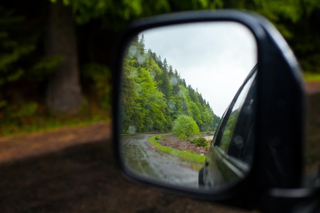 Reflacción de la carretera mojada y el bosque de verano en el espejo retrovisor lateral de un automóvil