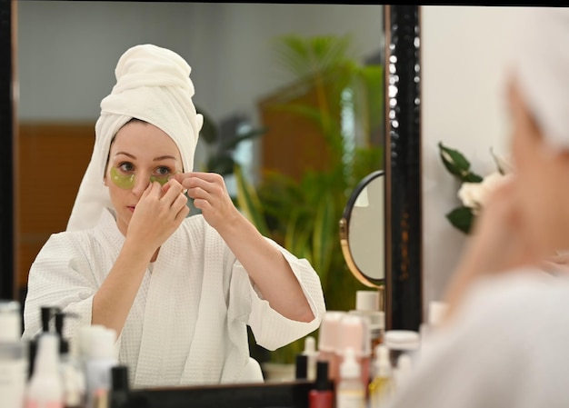 Reflejándose en el espejo de una hermosa mujer caucásica en bata de baño usando parches debajo de los ojos Autocuidado y rutina diaria de belleza