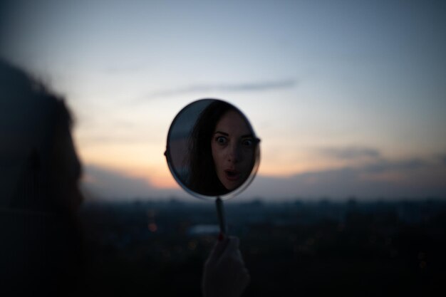 Foto reflexão de uma mulher chocada no espelho contra o céu durante o pôr do sol