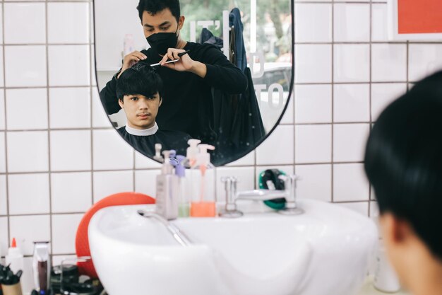 Reflexion aus dem Spiegel der jungen Leute im Friseursalon Friseur, der seinen Kunden die Haare kämmt