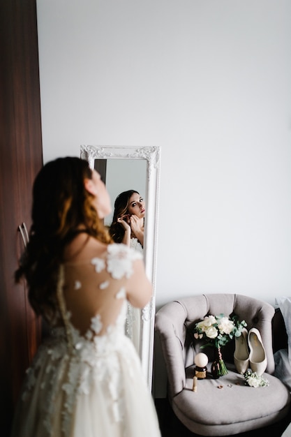 Reflexión en el espejo. La novia con un vestido blanco usa aretes de oro con perlas en la habitación cerca del espejo. En manos de la decoración de la novia. Mañana de bodas. Una mirada a la espalda de la mujer.