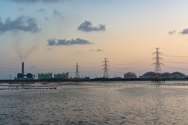 Reflexión del frente del río de la luz de la refinería de petróleo en el crepúsculo, fondo industrial del paisaje