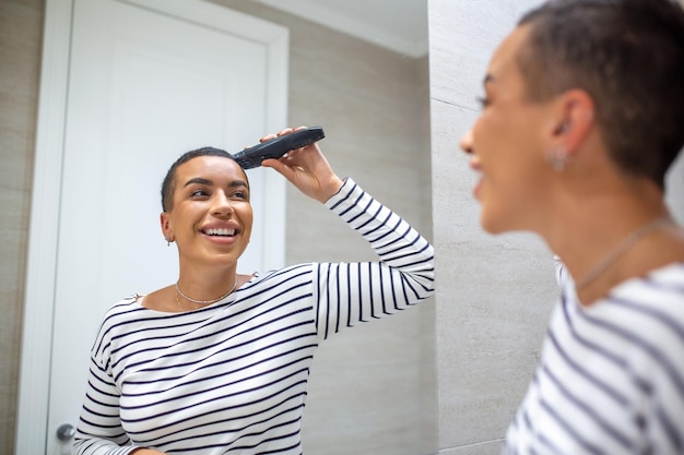 Reflexo de espelho de mulher de cabelos curtos em regata usando máquina de corte de cabelo