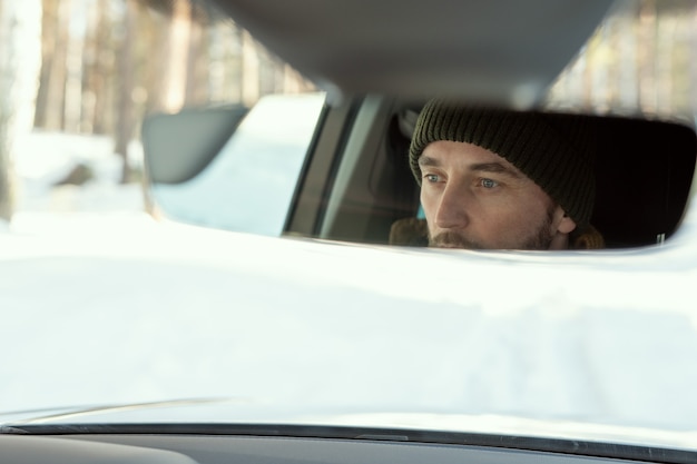 Reflexo do rosto de um jovem ativo em trajes de inverno, olhando pela janela da frente do carro enquanto dirige para a casa de campo ou algum outro lugar