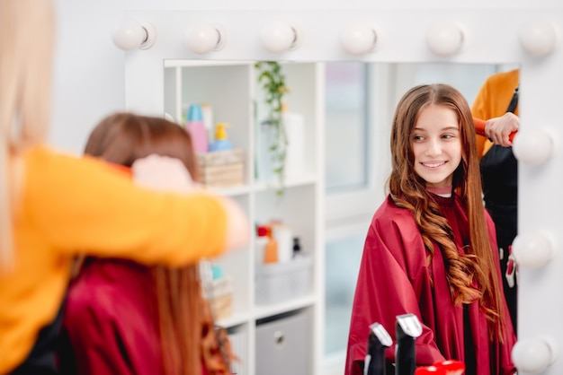 Reflexo no espelho de uma jovem e atraente modelo sorridente com cachos durante o processo de penteado no salão de beleza