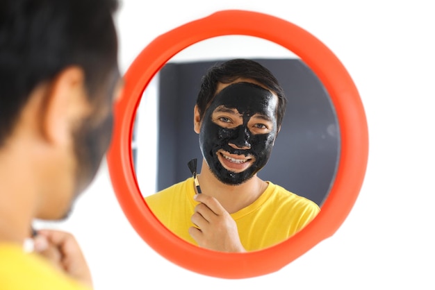 Reflexo no espelho do homem aplicando máscara de argila de carvão preto no rosto Conceito de beleza e cuidados com a pele