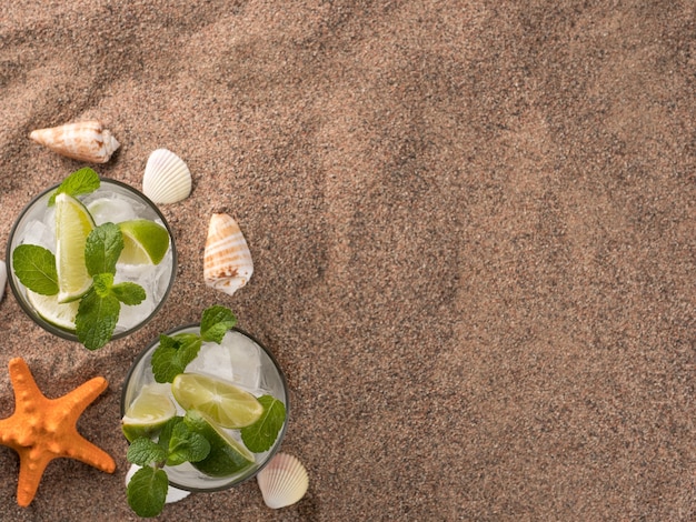 Una refrescante bebida de verano con mojito de lima y menta se encuentra en la arena con conchas marinas y estrellas de mar.