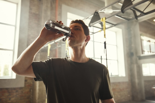 Refrescante. Joven atlético bebiendo agua fresca de la botella mientras hace ejercicio en el gimnasio