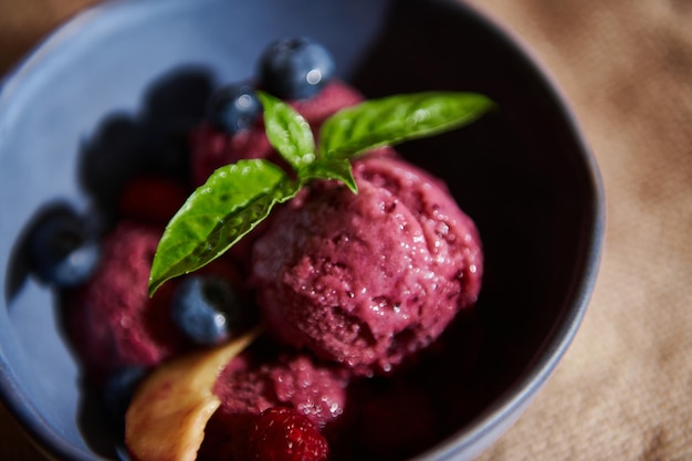 Refrescante sorbete de bayas moradas veganas crudas en tazón de cerámica azul marino con hojas de albahaca limón, arándanos y melocotones jugosos