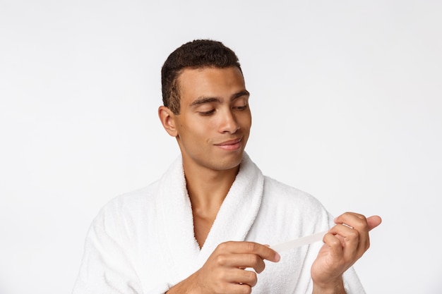 Refresque al hombre hermoso en la albornoz que hace la manicura para sí mismo con la lima de clavo aislada sobre blanco.