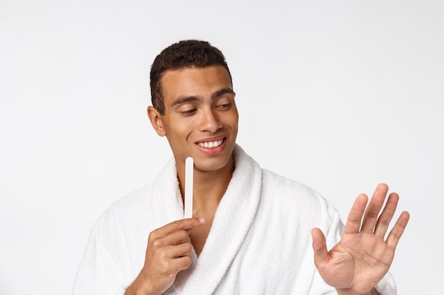 Refrigere o homem considerável no roupão que faz o manicure para si mesmo com a lixa de unha isolada sobre o branco.