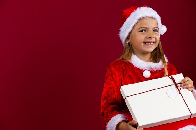 Regalo de apertura de niña. Retrato de una niña bonita con traje de Santa Claus y sonriendo. Pequeña bebé caucásica. Pared aislada roja.